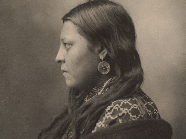 Side profile of a Native American wearing a pattered clothing and a dark shawl