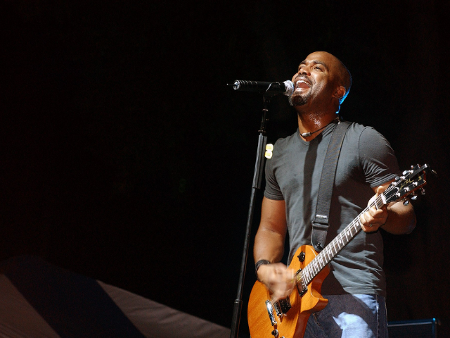 Darius Rucker wearing a grey t-shirt and jeans. He holds a brown guitar as he performs a song.