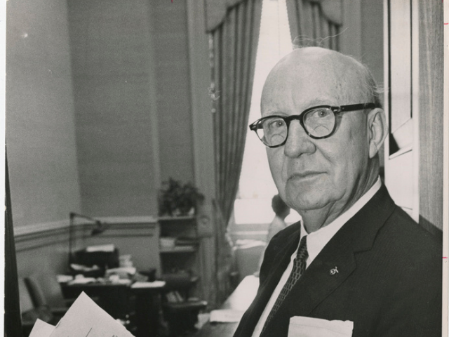 Edgar A. Brown wearing a dark suit and dark framed glasses holding some papers. 