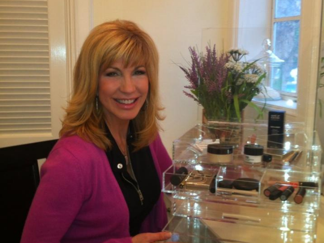 Leeza Gibbons smiling wearing a magenta sweater over black blouse next to a make-up stand.
