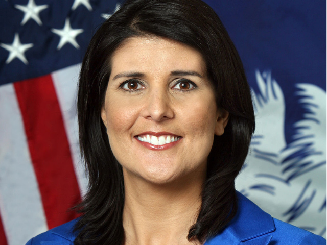 A smiling Nikki Haley wearing a royal blue blouse with the American flag and the South Carolina state flag in the background. 