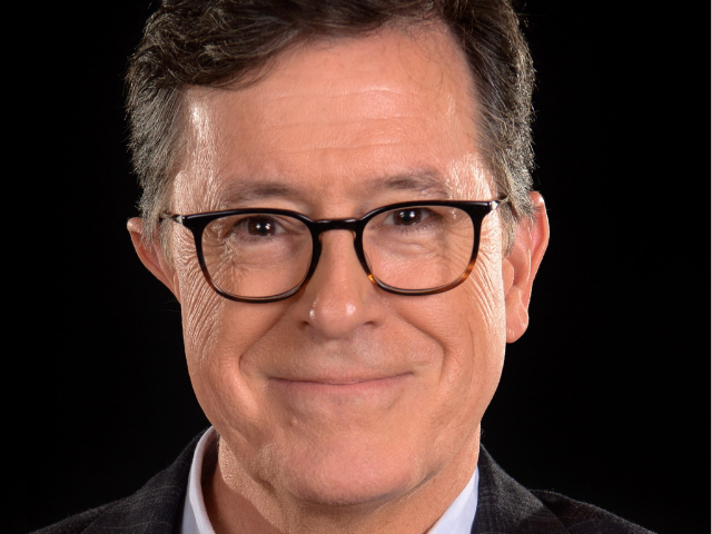 Stephen Colbert wearing dark rimmed glasses a dark sweater and light shirt. 
