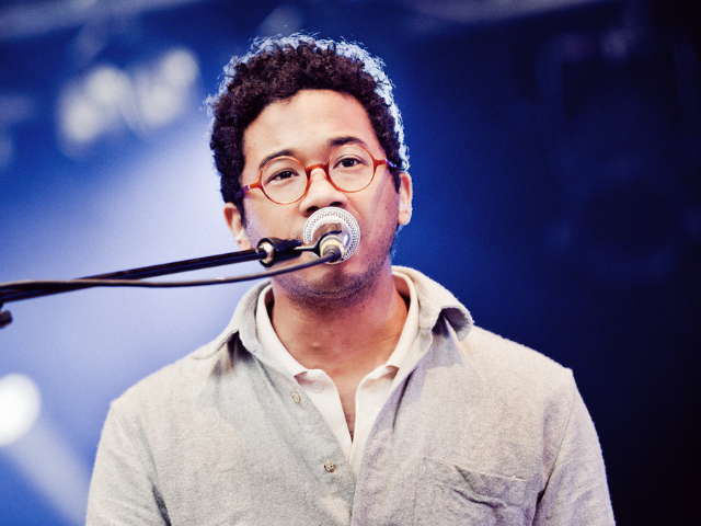 Toro y Moi wearing a light grey jacket singing into a silver and black microphone 