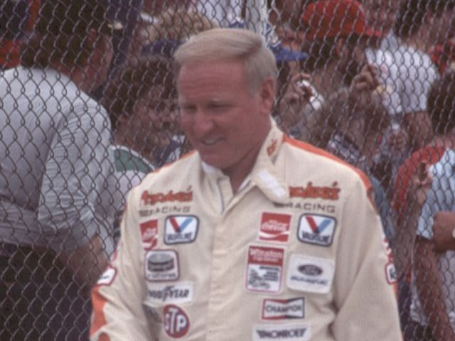 Cale Yarborough wearing a tan racing suit. Fans stand in the background behind a wired mesh fence. 