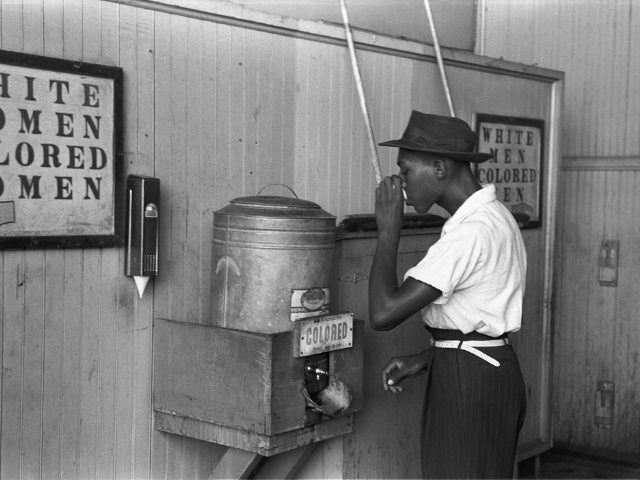 A black man drinking from a water cooler labelled Colored. 