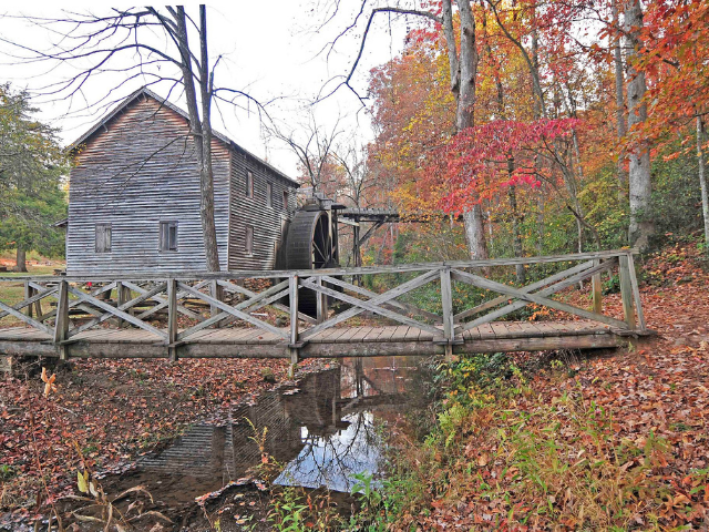 A wooden build with a mill wheel is next to a wooden bridge surround by trees.