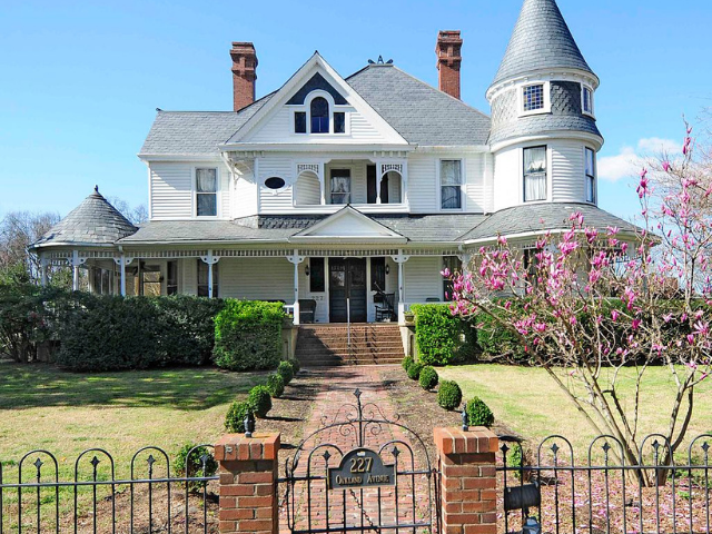 A stately white house with a gray roof, manicured lawn, and brick and iron fence.