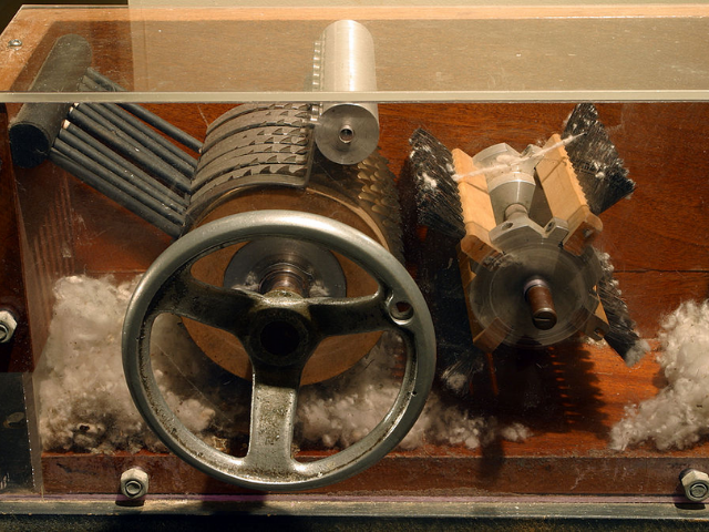 A machine of metal gears and wood behind a glass window with bits of cotton underneath. 