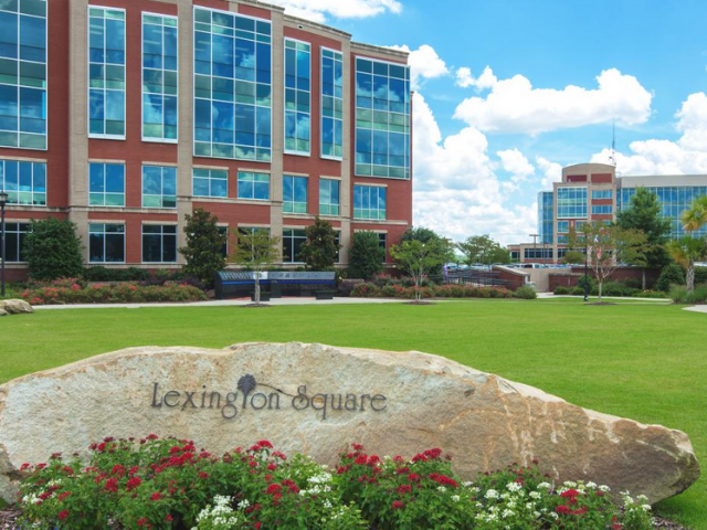 A greyish tan rock with the text Lexington Square sits in front of two brick and glass building and manicure landscaping. 