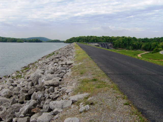 A dark gray road cuts between two grassy area. To left are rocks and a large body of water. 