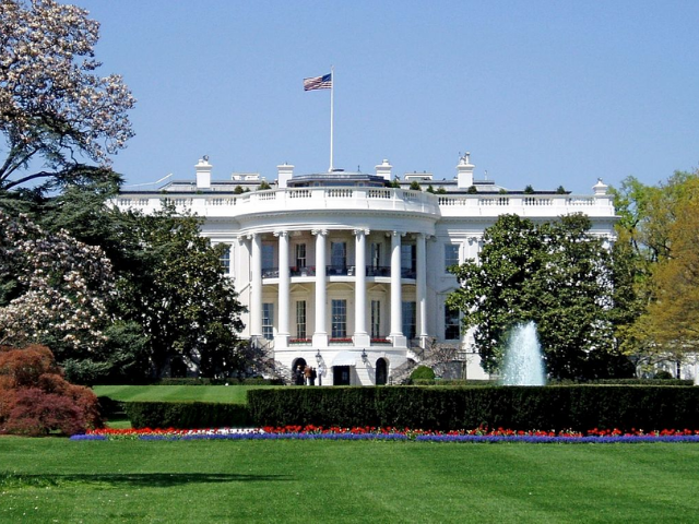A large white house with an American flag at the top of the roof surrounded by flowering trees and plants. 