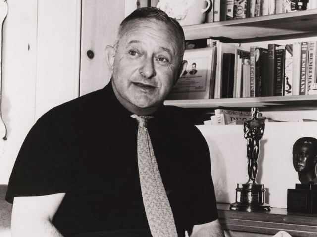 A man in a dark shirt and patterned tie stands next to a bookcase with an Oscar award.