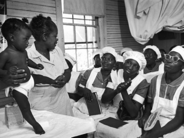 A standing woman holding a baby in front of a group of women.