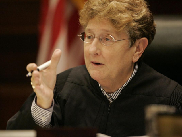 An older woman holding a pen and wearing a black judge robe.