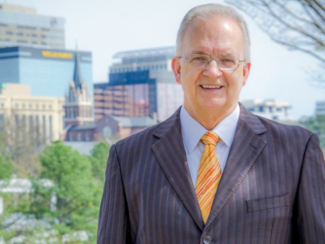 A older man in a navy blue suite with a yellow tie standing in front of a city