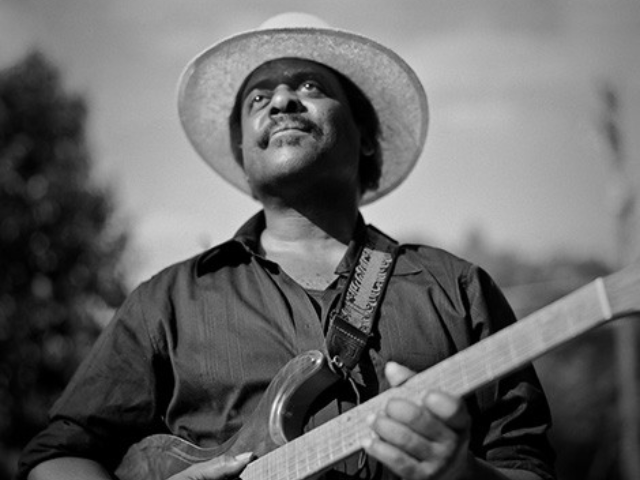 A middle-aged man wearing a wide brim hat and holding a guitar. 