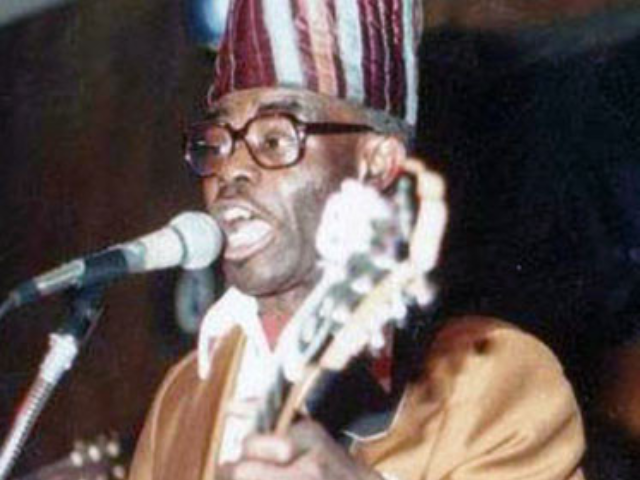 A middle aged man with a burgundy and white striped brimless hat and brown glasses sings into a microphone while holding a guitar. 